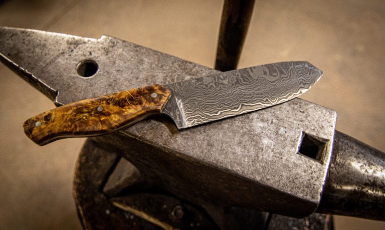 Handgeschmiedetes Damastmesser Ein Messer mit Holzgriff liegt auf einem Amboss.